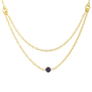 May include: A gold-colored, two-strand necklace with a small, round, dark purple gemstone pendant. The necklace has a delicate chain design and is displayed against a white background.