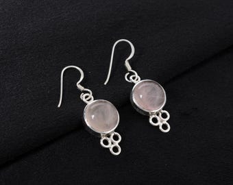 May include: A pair of silver earrings with rose quartz gemstones. Each earring features a round, pale pink gemstone set in a silver bezel. Below the gemstone is a decorative silver element with a cluster of small circles. The earrings hang from a curved silver hook.