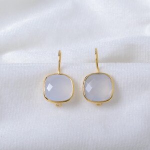 May include: A pair of gold-colored earrings with square, pale blue gemstones. The gemstones are faceted and set in a gold frame. The earrings hang from a simple gold hook. The background is a white, textured fabric.