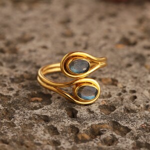Pu&ograve; includere: Un anello dorato con due pietre preziose ovali blu-grigie. Il design dell'anello presenta una doppia fascia che si curva attorno alle pietre preziose, creando un aspetto unico ed elegante. L'anello &egrave; posto su una superficie strutturata e neutra.
