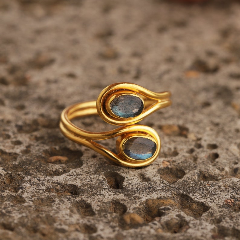 Pu&ograve; includere: Un anello dorato con due pietre preziose ovali blu-grigie. Il design dell'anello presenta una doppia fascia che si curva attorno alle pietre preziose, creando un aspetto unico ed elegante. L'anello &egrave; posto su una superficie strutturata e neutra.