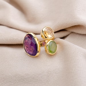 Op de afbeelding: Een gouden ring met drie edelstenen: een grote, gefacetteerde paarse amethist, een kleinere gele citrien en een groene peridot. De ring heeft een modern ontwerp met de edelstenen in gouden zettingen.