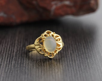 May include: A gold ring with an oval, light-colored gemstone. The ring has an ornate, openwork design around the gemstone. The ring is set against a dark wood surface, with a blurred, reddish-brown background.