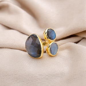 Può includere: Un anello dorato con tre pietre di labradorite. La pietra più grande ha una forma irregolare, mentre le altre due sono rotonde. Le pietre hanno un'iridescenza blu e grigia. L'anello è posto su uno sfondo di tessuto neutro.