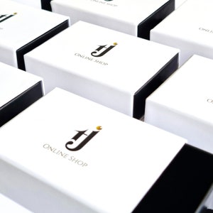 Pu&ograve; includere: Scatole regalo bianche con bordi neri disposte in una griglia. Ogni scatola presenta un logo con le lettere "TJ" e la scritta "ONLINE SHOP" in nero. Le scatole sono chiuse, indicando che sono pronte per l'imballaggio.