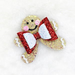 May include: Glittering gingerbread man with a red and white bow tie. The gingerbread man has pink cheeks and a smiling face. The design is on a white, fluffy background. This festive item is perfect for holiday decorations.