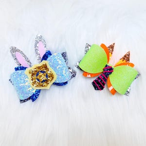 Peut inclure: Deux nœuds décoratifs pour cheveux. L'un présente des paillettes bleues avec des oreilles de lapin et un badge doré. L'autre a des accents verts, oranges et marron avec une cravate rayée. Les deux sont sur une surface blanche et duveteuse.