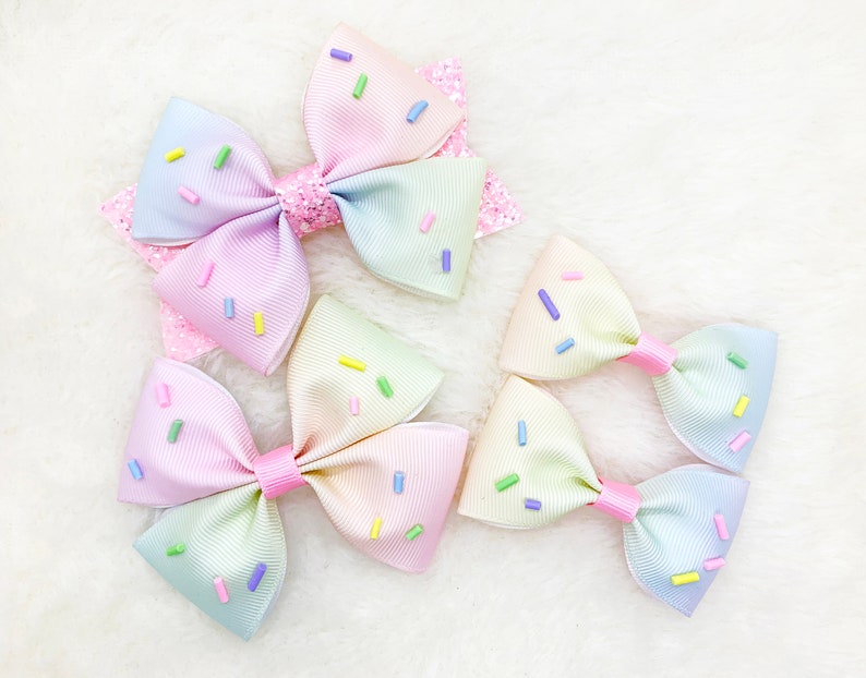 Pu&ograve; includere: Tre fiocchi per capelli arcobaleno pastello con un motivo a spolverata. I fiocchi sono fatti di nastro e hanno un anello centrale.