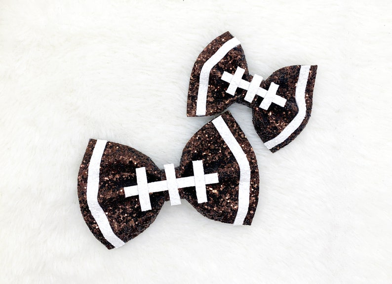 May include: Two brown glitter hair bows shaped like footballs with white stitching.