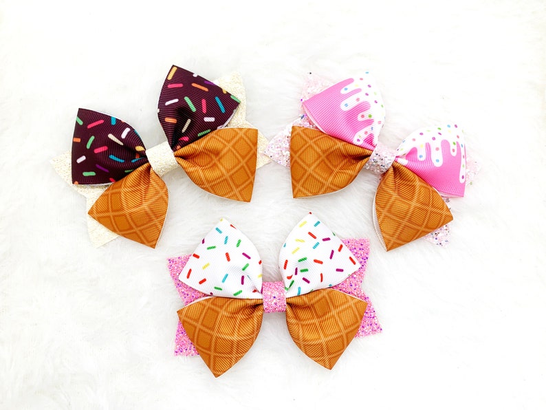 May include: Three handmade hair bows in the shape of ice cream cones. The bows are made of brown, pink, and white fabric with sprinkles and a waffle cone pattern.