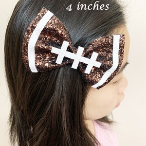 May include: A brown and white glitter football-shaped hair bow with white stitching. The bow is 4 inches wide.