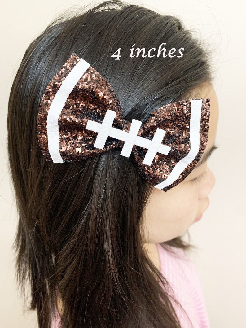 May include: A brown and white glitter football-shaped hair bow with white stitching. The bow is 4 inches wide.