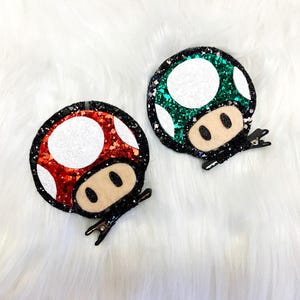May include: Two mushroom-shaped hair clips. One is red with white spots, the other is green with white spots. Both have a black border and a tan face with black eyes. The clips are on a white, fluffy surface.