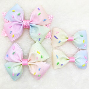Pu&ograve; includere: Tre fiocchi per capelli in ombr&eacute; arcobaleno pastello con codette colorate. I fiocchi sono realizzati in nastro e sono annodati. I fiocchi sono disposti in fila dal pi&ugrave; grande al pi&ugrave; piccolo.