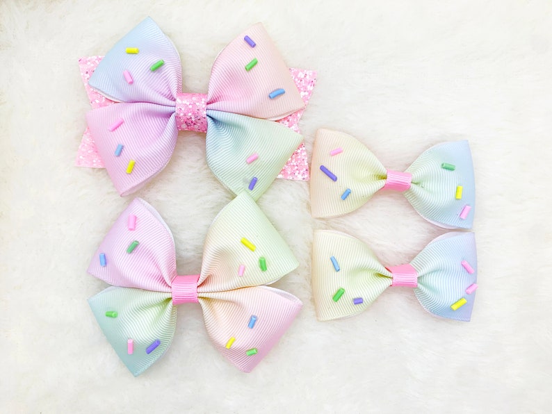 Pu&ograve; includere: Tre fiocchi per capelli in ombr&eacute; arcobaleno pastello con codette colorate. I fiocchi sono realizzati in nastro e sono annodati. I fiocchi sono disposti in fila dal pi&ugrave; grande al pi&ugrave; piccolo.