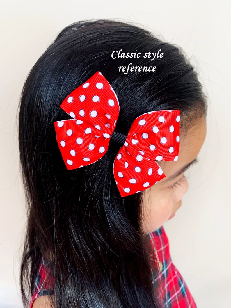 May include: A red and white polka dot bow with a black elastic band. The bow is tied in a classic style and is worn in a girl's hair.