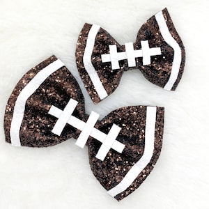 May include: Two brown and white glitter hair bows shaped like footballs. The bows have white stitching details.