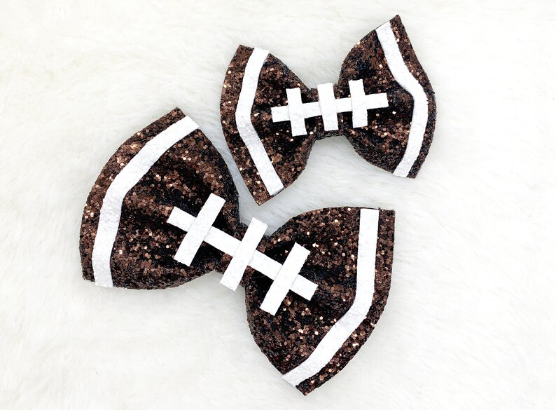 May include: Two brown and white glitter hair bows shaped like footballs. The bows have white stitching details.