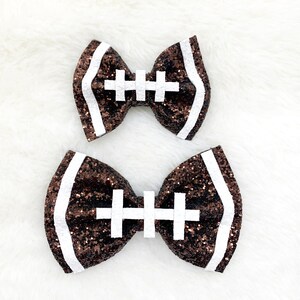 May include: Two brown and white glitter football-shaped hair bows. The bows are tied with a white ribbon.