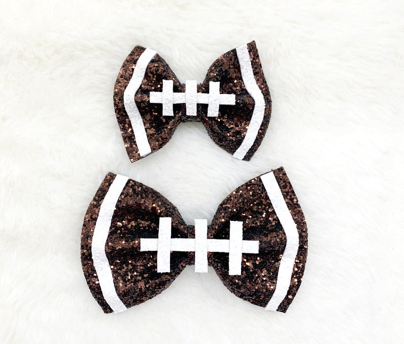 May include: Two brown and white glitter football-shaped hair bows. The bows are tied with a white ribbon.