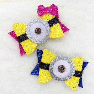 May include: Two yellow, blue and pink glitter hair bows with silver and white eyes. The bows are shaped like a minion from the film Despicable Me.