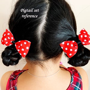 Pu&ograve; includere: Una bambina con i capelli neri indossa un vestito a quadri rossi e neri e ha due fiocchi rossi con pois bianchi nelle sue trecce. Il testo "Pigtail set reference" &egrave; scritto sopra la sua testa.