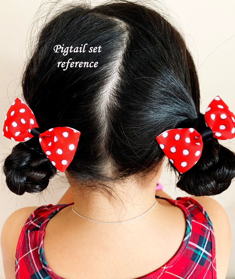 Pu&ograve; includere: Una bambina con i capelli neri indossa un vestito a quadri rossi e neri e ha due fiocchi rossi con pois bianchi nelle sue trecce. Il testo "Pigtail set reference" &egrave; scritto sopra la sua testa.
