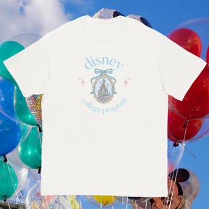 May include: White t-shirt with a light blue design that reads "disney college program". The design features a castle illustration within a decorative frame, a bow, and star accents. The background includes colourful balloons.