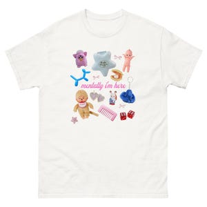 Peut inclure: T-shirt blanc orné d'une collection de petits jouets et accessoires. Le motif comprend un Furby rose, un animal ballon bleu, une poupée, un peigne, des dés et le texte "mentally I'm here".