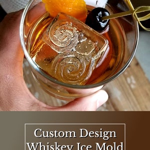 May include: A close-up of a whiskey old fashioned cocktail with a large ice cube, an orange peel garnish, and a maraschino cherry. The ice cube has a swirling design. The text "Custom Design Whiskey Ice Mold" and "Speakeasy Ice Molds" is visible below the image.
