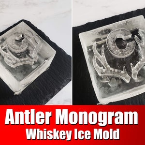 May include: Clear ice mold with an antler monogram design. The ice cube is square-shaped and sits on a black surface. The text "Antler Monogram Whiskey Ice Mold" is displayed in white letters on a red background.