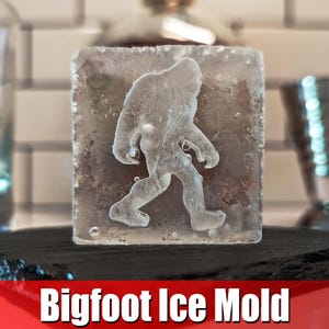 May include: A clear ice mold with a raised design of Bigfoot walking. The mold is on a black slate base.