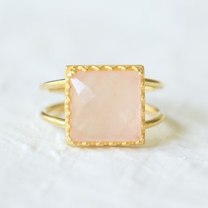 May include: A gold-colored ring with a square, light pink gemstone. The gemstone is set in a textured gold frame and sits atop a double-banded ring. The ring is displayed on a white, textured surface.