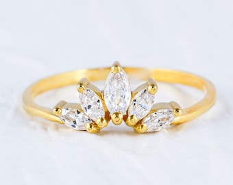 May include: A gold-colored ring with a crown-like design. The ring features five clear, marquise-cut gemstones set in a symmetrical pattern. The band is thin and simple, and the ring is displayed on a white surface.