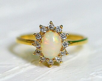 May include: A gold ring featuring an oval opal gemstone surrounded by a halo of small, clear crystals. The ring's band is thin and gold, and the setting is a starburst design. The ring is set against a white background.