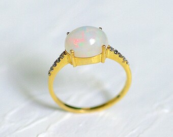 May include: A gold ring featuring a large, oval opal gemstone. The opal has a milky white base with flashes of color. The band is gold with small, dark gemstones set along the sides of the opal.