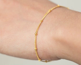 May include: A delicate gold bracelet featuring a fine chain interspersed with small, evenly spaced beads. The bracelet is a warm, bright gold color and appears to be worn on a wrist. The design is simple and elegant, suitable for everyday wear.