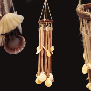 Pode incluir: Um móbile de vento de madeira com conchas penduradas nele. O móbile é feito de materiais naturais e tem um visual rústico. As conchas são de cor bege claro e estão dispostas em um cacho na parte inferior do móbile.