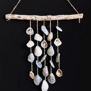 Può includere: Un mobile in legno di deriva e conchiglie con diverse conchiglie di ostriche appese a un pezzo di legno di deriva. Le conchiglie sono di varie tonalità di grigio e bianco.