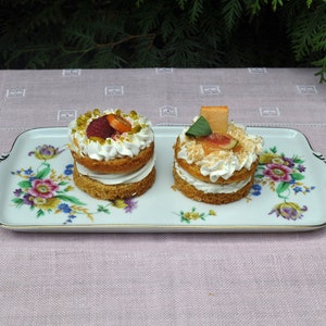 May include: Two small cakes on a white plate with floral designs. The cakes are topped with whipped cream and fruit. One cake has raspberries and orange slices, the other has figs and a slice of orange.
