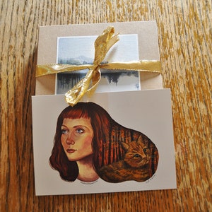 May include: A greeting card with a painting of a woman with red hair and a cat in a forest setting. The card is tied with a gold ribbon.