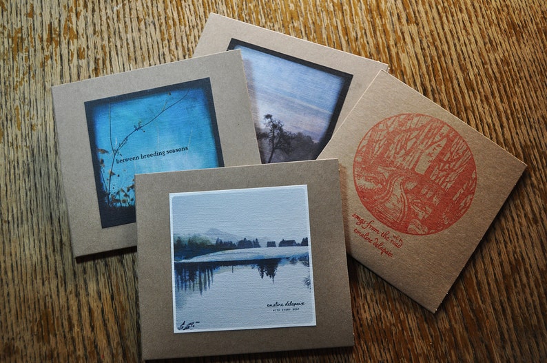 May include: Four brown paper CD covers with different images and text. The covers are stacked on top of each other. The top cover has a blue and white image with the text "between breeding seasons". The second cover has a blue and white image of a lake with the text "winter solstice". The third cover has a blue and white image of a forest with the text "songs from the road". The bottom cover has a red and white image of a tree with the text "creative deluge".