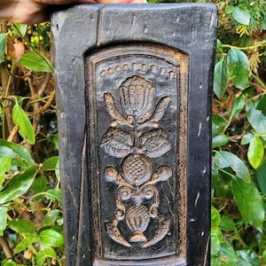 May include: A rectangular black wooden mold featuring a deeply carved relief design of a central floral motif with leaves and a decorative base. The wood has a weathered, dark finish with visible grain and natural imperfections throughout the surface.