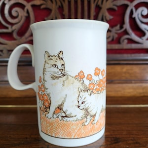May include: White ceramic mug with a cat and kitten illustration and orange flowers. The mug has a curved handle and a straight rim. The cat illustration is in shades of brown and white.