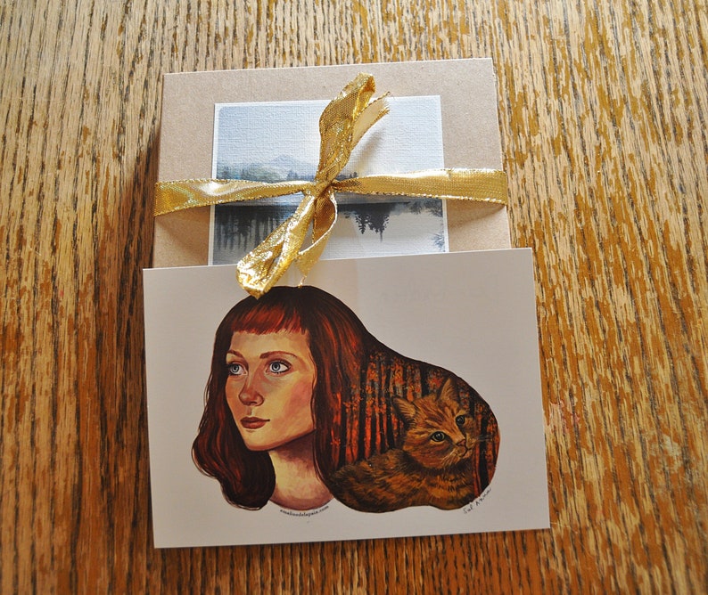 May include: A greeting card with a painting of a woman with red hair and a cat in a forest setting. The card is tied with a gold ribbon.