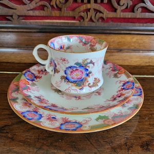 May include: A white porcelain teacup and saucer set with a floral pattern in shades of pink, blue, and green. The cup has a gold rim and the saucer has a pink rim.