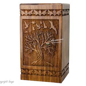 Puede incluir: Una urna de cremación de madera con un diseño tallado de árbol de la vida y pájaros volando a su alrededor. La urna está hecha de madera oscura y tiene una forma rectangular.