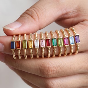 Peut inclure: Une collection de douze bagues dorées, chacune ornée d'une pierre précieuse rectangulaire taille baguette de couleur différente, incluant bleu, orange, vert, jaune, transparent, rose, rouge, émeraude, lime, magenta, bleu clair et violet, sur un anneau torsadé.