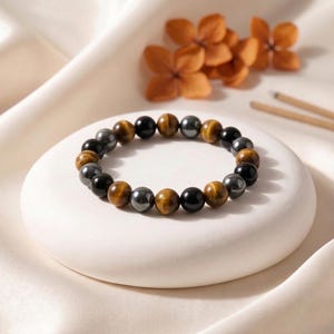May include: A beaded bracelet featuring a mix of black, brown, and gray spherical beads. The bracelet is displayed on a white, circular platform, with dried orange flowers and wooden sticks in the background.