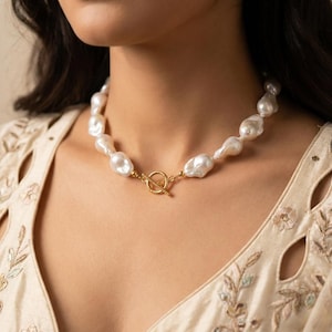 Pode incluir: Um colar com pérolas barrocas brancas irregulares enfiadas, finalizado com um fecho de metal dourado em forma de T posicionado no centro. As pérolas variam em forma e tamanho, criando uma aparência texturizada ao redor do pescoço.